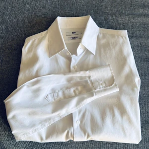 Vit skjorta från Uniqlo - Klassisk vit skjorta från Uniqlo med lång ärm och knappar framtill. Skjortan har en stilren design med normal passform och är tillverkad i bomull. Behöver ej strykas. Perfekt till både vardags och mer fina tillfällen tex studenten. Hör av er vid intresse/ frågor!