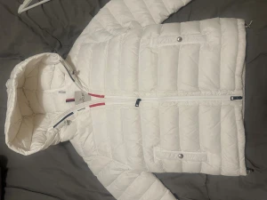 Vit dunjacka från Moncler med huva - Snygg vit dunjacka från Moncler med huva och quiltad design. Jackan har dragkedja framtill, två fickor med knappstängning och Moncler-logga på ärmen. Insidan har klassisk Moncler-etikett och detaljer i rött och blått vid dragkedjan. Perfekt för kalla dagar.
