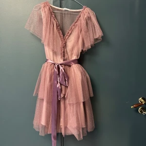 Ida Sjöstedt klänning strl 38 - Superfin rosa kortklänning från Ida Sjöstedt med glittrigt tylltyg, volanger och v-ringning. Klänningen har lager på lager-kjol, korta ärmar och ett sidenband i midjan som knyts till rosett. Perfekt för dig som vill sticka ut med en romantisk look.