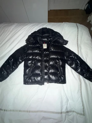Svart pufferjacka från Moncler - Säljer en svart pufferjacka från Moncler med glansig finish och dunfyllning. Jackan har huva, dragkedja framtill, långa ärmar och Moncler-logga på ärmen. Flera fickor med dragkedja och knappdetaljer. Perfekt för kalla dagar och riktigt snygg streetstil. Väldigt bra skick utan defekter!❤️❤️