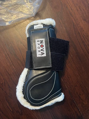 Nova Sport svarta benskydd pony - ALDRIG ANVÄNDA! Säljer ett par svarta benskydd från Nova Sport i storlek pony. Utsidan är i svart syntetmaterial med vita kontrastsömmar och insidan är fodrad med mjuk, vit fuskpäls. Breda kardborreband för enkel justering och bra passform. 