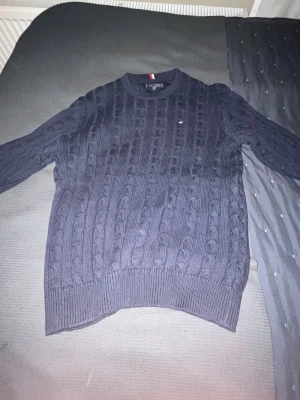 Mörkblå stickad tröja Tommy Hilfiger - Snygg mörkblå stickad tröja från Tommy Hilfiger med klassiskt kabelstickat mönster. Rund hals och långa ärmar, liten logga på bröstet. Perfekt för dig som gillar stilrena och tidlösa plagg med en sportig touch.