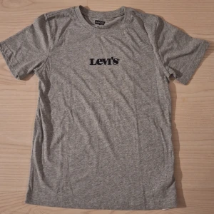 Ny grå Levi's t-shirt stl 14 år - Snygg grå t-shirt från Levi's med klassisk svart broderad logga på bröstet. T-shirten har rund hals och korta ärmar, tillverkad i mjuk bomull som är skön mot huden. Perfekt för dig som gillar enkel och stilren design.
