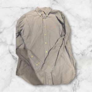 Rutig skjorta från Ralph Lauren - Snygg rutig skjorta från Ralph Lauren i ljus blå  och grå toner med klassisk krage och långa ärmar. Skjortan har gula knappar framtill och en liten röd broderad logga på bröstet. Perfekt för dig som gillar stilrena detaljer och tidlös design.