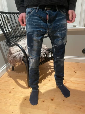 Dsquared2 blå jeans med slitningar - Säljer ett par blå jeans från Dsquared2 i storlek32 bredd 34 längd . Jeansen har coola slitningar, färgstänk och en snygg tvättad look. De har klassisk femficksdesign, smal passform och är tillverkade i jeansmaterial med detaljerade sömmar. Perfekt för dig som gillar streetstyle.