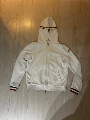 Vit Moncler hoodie med ränder - Vit hoodie från Moncler med hel dragkedja, huva och fickor framtill. Ränder i rött, blått och svart på huvan och ärmsluten ger en sportig vibe. Moncler-logga på vänster ärm. Mjuk bomullskvalitet, perfekt för chill dagar. Finns en liten fläck men det går säkert att ta bort i tvätten.                  Pris kan diskutera vid snabb affär!!!!
