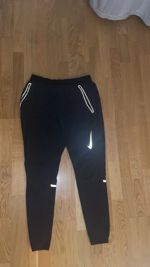 Svarta Nike träningsbyxor med reflex - Svarta träningsbyxor från Nike med smal passform och snygga reflexdetaljer vid fickor, ben och logga. Byxorna har elastisk midja och är perfekta för löpning eller gym. Materialet är mjukt och stretchigt för maximal komfort.