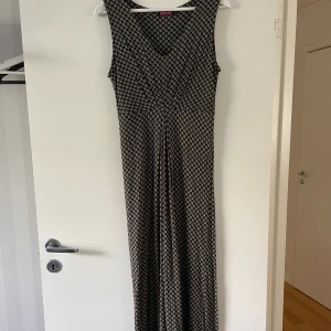Svart långklänning Indiska mönstrad S - Långklänning från Indiska i svart med små beige mönster. Ärmlös modell med v-ringning och omlott-effekt upptill. Tunt, mjukt tyg som faller snyggt. Perfekt för dig som gillar boho-vibe och vill ha något bekvämt men ändå stilrent.