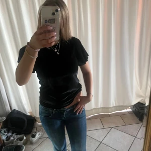 Svart sammetsblus med volangärm - Svart blus i mjuk sammet med rund halsringning och korta volangärmar. Snygg passform som sitter lite tightare över kroppen och ger en elegant look. Perfekt att styla med jeans eller kjol för en trendig outfit.