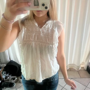 Vit blus med volang och spetsdetaljer - Supersöt vit blus i luftig bomull med volangärmar och fina spetsdetaljer över bröstet. Blusen har v-ringning och rynkade partier som ger en romantisk vibe. Perfekt till jeans eller kjol för en fräsch look i sommar.