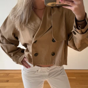 Kort beige trenchcoat med knappar - Säljer en snygg croppad trenchcoat i beige med dubbelknäppning och stora svarta knappar. Jackan har klassisk krage, långa ärmar och är tillverkad i bomull. Perfekt för dig som gillar trendiga och stilrena plagg.