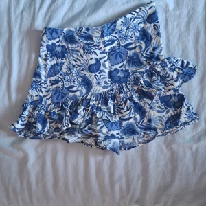 Blommiga volangshorts från Zara XS - Supersöta shorts från Zara i storlek XS med blått och vitt blommönster. De har hög midja och volangdetaljer längs benen. Shortsen ser lite ut som en kjol fram i från.