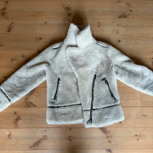 Vit teddyjacka med dragkedjor - Säljer en fluffig vit teddyjacka med svarta detaljer och dragkedjor på fickorna. Jackan har hög krage och markerade sömmar som ger en cool look. Perfekt för dig som vill ha en mysig men trendig jacka till höst och vinter.