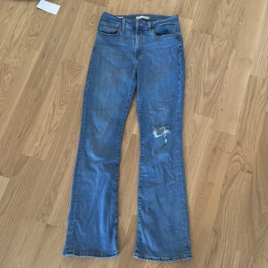 Levi's 725 High Rise Bootcut Jeans i strl 28 (S) - Säljer ett par blå Levi's 725 High Rise Bootcut jeans i klassisk denim. De har hög midja, bootcut-ben och en snygg slitning på ena benet. Perfekta för dig som gillar en tidlös look med lite edge. Klassiska femficksjeans med Levi's läderpatch bak.