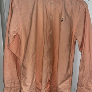 Persikofärgad skjorta från Ralph Lauren - Snygg persikofärgad skjorta från Ralph Lauren med klassisk krage och knappar framtill. Skjortan har lång ärm och ikonisk broderad logga på bröstet. Tillverkad i 100% bomull för en skön och luftig känsla.