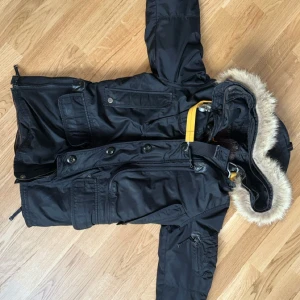 Svart vinterjacka Parajumpers med päls - Säljer en svart Parajumpers vinterjacka med avtagbar huva och fuskpäls. Jackan har flera fickor, quiltat foder och ikonisk gul rem vid kragen. Perfekt för kalla dagar och riktigt snygg streetstil.