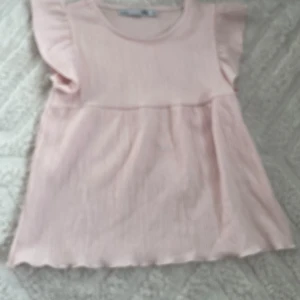 Ljusrosa blus med volangärm från lager157 - Supersöt ljusrosa blus från lager157 i storlek 140. Blusen har volangärmar och är tillverkad i ett ribbat, mjukt syntetmaterial. Den har en rund halsringning och ett flowigt fall med lätt struktur i tyget. Perfekt för dig som gillar gulliga och feminina toppar.