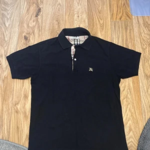 Svart pikétröja från Burberry - Klassisk svart pikétröja från Burberry med ikoniskt rutigt tyg på insidan av kragen och knappslån. Tröjan har korta ärmar, broderad logga på bröstet och snygg passform. Perfekt för dig som gillar stilrena och tidlösa plagg.