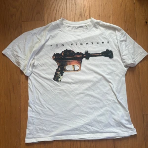 Vit Foo Fighters t-shirt loose fit - Vit t-shirt från Foo Fighters med coolt tryck av en retro raygun på bröstet och bandets logga ovanför. Modellen är loose fit och har korta ärmar. Perfekt för dig som gillar bandmerch och vill ha en avslappnad stil. Priset kan självklart diskuteras 