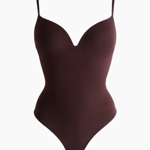 Brun tight bodysuit med smala axelband - Säljer en mörk röd, tight body med smala justerbara axelband och djup v-ringning. Bodysuiten har en slät, figurnära passform och är tillverkad i ett stretchigt syntetmaterial. Perfekt att styla med jeans eller kjol för en trendig look.
