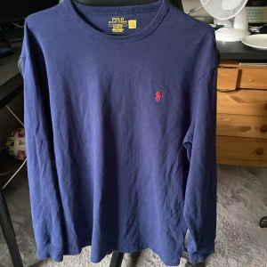 Blå långärmad tröja från Polo Ralph Lauren - Säljer en klassisk marinblå långärmad tröja från Polo Ralph Lauren med rund hals och röd broderad logga på bröstet. Tröjan är i bomull och har en relaxed fit som passar perfekt till jeans eller chinos. Nypris 1300