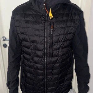 Svart pufferjacka från Parajumpers - Svart pufferjacka från Parajumpers med huva, quiltad framsida och rygg samt släta ärmar. Jackan har dragkedja framtill, bröstficka med dragkedja och den ikoniska gula detaljen vid halsen. Perfekt för kyliga dagar och snygg streetstil.
