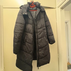 Svart lång pufferjacka från Pieces - Lång svart pufferjacka från Pieces med huva och dragkedja framtill. Använd några gånger. 