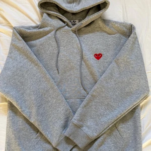 Grå hoodie Comme des Garçons Play - Grå hoodie från Comme des Garçons Play med klassiskt rött hjärta med ögon broderat på bröstet. Tröjan har huva med snörning, känguruficka och långa ärmar. Perfekt för dig som gillar stilrena streetwear-plagg med en twist.
