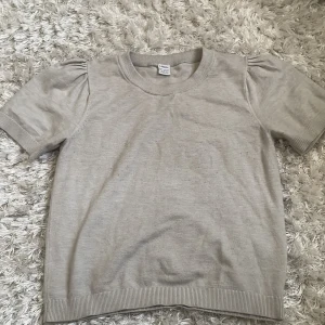 Beige kortärmad tröja från Lindex - Snygg beige tröja från Lindex med kort puffärm och ribbade muddar vid hals, ärmslut och nederkant. Enkel och stilren design som passar perfekt till jeans eller kjol. Mjuk och skön bomullskvalitet.