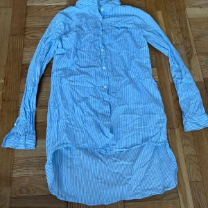 Blåvit randig skjorta från Name It - Snygg långärmad skjorta från Name It i blått och vitt med smala ränder. Skjortan har klassisk krage, knappar framtill och längre bakstycke för en cool look. Perfekt för dig som gillar stilrena och fräscha plagg.