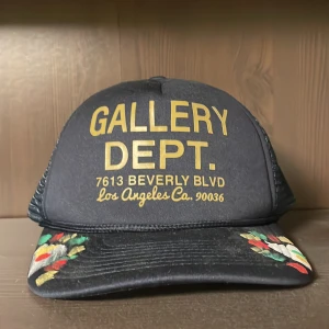 Gallery Dept keps - Blå truckerkeps från Gallery Dept. med gult tryck framtill och handmålade detaljer i rött, grönt, vitt och gult på skärmen. Kepsen har mesh på sidorna och justerbar passform. Perfekt statement-piece för dig som vill sticka ut.