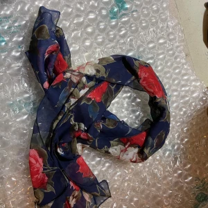 Blommig mörkblå sjal i silke-look - Snygg och tunn halsduk med blommigt mönster i rött, vitt och grönt på en mörkblå botten. Perfekt accessoar för att lyfta din outfit med lite färg och stil. Lätt och luftig, passar bra att knyta runt halsen eller i håret.