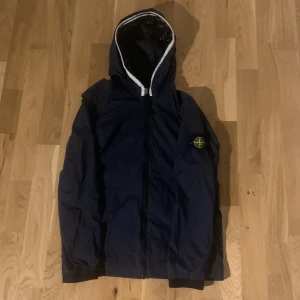Mörkblå vindjacka från Stone Island - Snygg mörkblå vindjacka från Stone Island med huva och klassisk logotyp på armen. Nypris för jackan är 2049kr. Jackan är i junior storlek som passar för barn 12-13 år. 158-166cm. Använd bara några få gånger i den bästa skicket någonsin! 