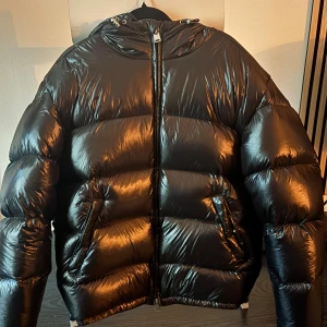 Svart pufferjacka från ZARA - Säljer en svart, glansig pufferjacka från ZARA i storlek M. Jackan har huva, dragkedja framtill och två fickor med knapp. Perfekt för kalla dagar när du vill hålla värmen och ändå se cool ut. Den trändiga jackan är knappt använd. 