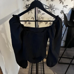 Svart blus med puffärmar - Svart blus med smockad, tight kropp och stora puffiga långärmar. Toppen har en offshoulder-design och är tillverkad i ett lätt, strukturerat tyg som ger en cool och trendig look. Perfekt för dig som gillar statement-ärmar och vill sticka ut.