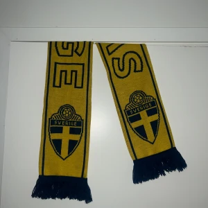 Sverige halsduk i gult och blått - Gul och blå halsduk med Sveriges landslagssköld och texten 'Sverige' i stora bokstäver. Stickad modell med blå fransar i ändarna, perfekt för att visa ditt stöd på matchen eller när du vill representera Sverige.