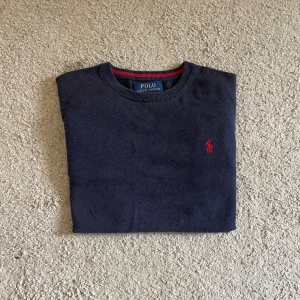 Mörkblå tröja från Ralph Lauren - Mörkblå tröja från Polo Ralph Lauren i mjuk pima bomull. Klassisk rund halsringning, ribbade muddar och röd broderad logga på bröstet. Perfekt för dig som gillar stilrena och tidlösa plagg.