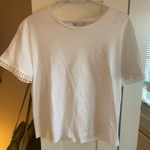 Vit T-shirt med detalj från Zara!  - Vit topp från Zara i bomull med rund halsringning och korta ärmar. Ärmsluten har snygga spetsdetaljer i ett rutigt mönster som ger en extra touch (porto tillkommer). 