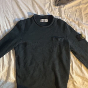 Mörkgrön stickad tröja Stone Island - Mörkgrön stickad tröja från Stone Island med klassisk patch på ärmen och en liten bröstficka. Tröjan har rund halsringning och långa ärmar, perfekt för dig som gillar stilren och clean design.