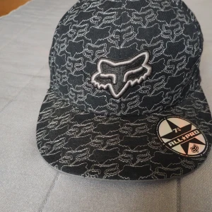 Svart Fox keps med broderat mönster - Snygg svart keps från Fox med broderat logomönster över hela kepsen och stor Fox-logga framtill. Tillverkad i 100% ull och har en klassisk rak skärm. Perfekt för dig som gillar streetwear och vill sticka ut med ett coolt mönster.