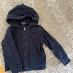 Marinblå hoodie från Polo Ralph Lauren - Mysig marinblå hoodie från Polo Ralph Lauren med dragkedja och huva. Tröjan har det klassiska röda Polo-logot broderat på bröstet och praktiska fickor framtill. Perfekt för en chill och avslappnad stil.