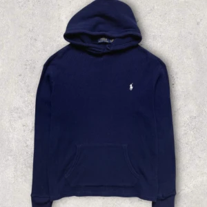 Marinblå hoodie från Polo Ralph Lauren - Snygg marinblå hoodie från Polo Ralph Lauren med klassisk broderad logga på bröstet. Tröjan är tillverkad i ett strukturerat bomullstyg, har huva och känguruficka framtill. Perfekt för en chill och stilren look.
