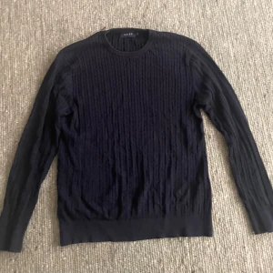 Mörkblå kabelstickad tröja Riley - Kabelstickad mörkblå tröja från Riley med rund halsringning och ribbade muddar. Tröjan har lång ärm och klassisk passform, perfekt för lager på lager under kyliga dagar. Snygg struktur i materialet som ger en stilren look.   NYPRIS: 900kr