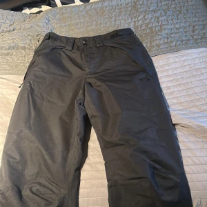 Svarta täckbyxor från Oakley XS - Svarta täckbyxor från Oakley i performance fit. Byxorna har dragkedjor vid fickorna och bälteshällor i midjan. Perfekta för vinterns aktiviteter och håller dig både torr och bekväm. Snygg och stilren design med Oakley-logga på benet.