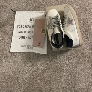 Golden Goose Superstar sneakers vita/blå - Golden Goose Superstar sneakers i vitt skinn med grå mockadetaljer och blå häl. Klassisk stjärndesign på sidan, grå snörning och platt sula. Snygg och trendig modell med cool vintagekänsla. Levereras med originalpåse och tillbehör.