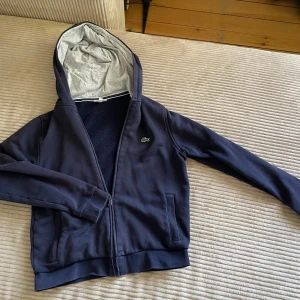 Mörkblå hoodie från Lacoste - Mörkblå hoodie från Lacoste med dragkedja och två fickor framtill. Tröjan har en grå insida i huvan och den klassiska krokodilloggan på bröstet. Perfekt för en avslappnad och sportig stil.
