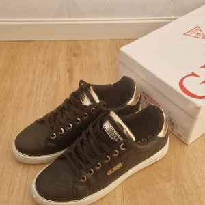 Svarta sneakers från Guess med gulddetaljer - Snygga svarta sneakers från Guess i storlek 38 med guldfärgade detaljer och logga på sidan. Skorna har snörning, vit platt sula och diskret mönster i materialet. Perfekta för dig som vill ha en stilren men ändå lyxig look.