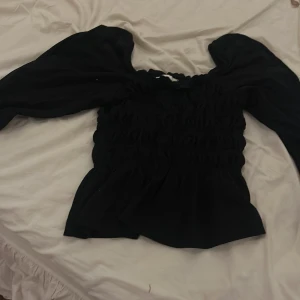 Svart blus med puffärmar från H&M - Säljer en svart blus från H&M i storlek XS. Toppen har smockad kropp och stora puffiga långärmar med resår vid axlarna. Den har en feminin och trendig look, perfekt för dig som gillar statement-ärmar och en figurnära passform över bålen.