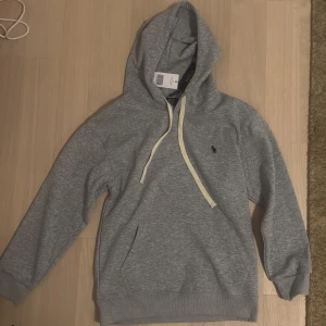 Grå hoodie från Polo Ralph Lauren - Snygg grå hoodie från Polo Ralph Lauren med klassisk huva och vita snören. Tröjan har en broderad logga på bröstet och en stor magficka. Perfekt för en chill och stilren look. Tillverkad i mjuk bomull för extra komfort. Den passar mer åt XS hållet‼️NYRPIS: 1951kr