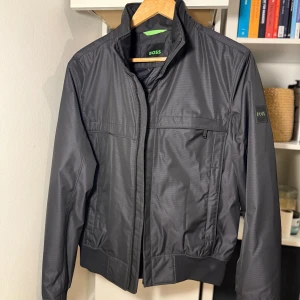 Svart vindjacka från BOSS - Snygg svart vindjacka från BOSS med hög krage, dragkedja och diskret logga på ärmen. Jackan har ribbade muddar och en minimalistisk design med fickor framtill. Perfekt för dig som gillar stilren och sportig look.